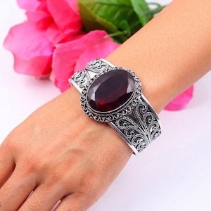May include: A silver cuff bracelet featuring an oval-shaped, deep red faceted gemstone at the centre. The metal band is decorated with intricate, swirling filigree patterns that extend from the central stone setting across the entire cuff.