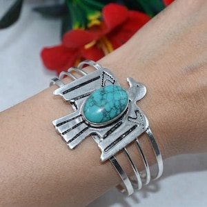 May include: A silver-toned cuff bracelet featuring a bird-shaped centrepiece with an oval turquoise stone. The metal bird has etched line details on its wings and tail, and the band consists of three thin, parallel silver-toned metal wires.