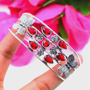 Puede incluir: Una pulsera de brazalete de plata con dos motivos de mariposa en los extremos, decorada con seis piedras rojas en forma de pera y dos pequeñas piedras rojas redondas, intercaladas con cuatro pequeños detalles de flores de plata.