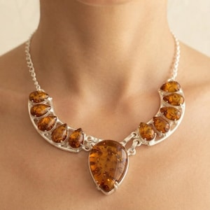 May include: A silver necklace featuring a large, central, pear-shaped amber stone surrounded by smaller, teardrop-shaped amber stones set in silver frames. The necklace is connected to a delicate silver chain that rests around the neck.