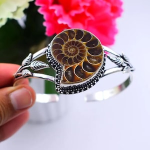 Puede incluir: Una pulsera de plata tipo brazalete con un fósil de amonita en espiral de color marrón y bronce en el centro. La banda metálica está decorada con motivos de hojas detallados a ambos lados y el engaste tiene un borde perlado alrededor.