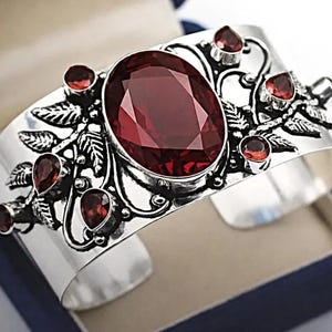 May include: A silver cuff bracelet featuring a large oval-cut dark red gemstone in the centre, surrounded by smaller pear-shaped and round red stones. The metal band is decorated with intricate embossed leaf patterns and swirling metal wire details.