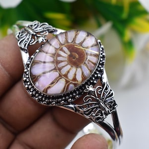 May include: A silver-toned cuff bracelet featuring a central oval-shaped ammonite fossil with brown and pink spiral patterns. The metal band is decorated with intricate butterfly motifs on both sides of the fossil and a beaded border.