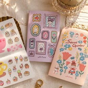 May include: A set of three notebooks and sticker sheets. One notebook is light purple with framed illustrations of a rabbit, a person, and a cake. Another is light pink with floral patterns and the text Secret Garden. A sheet features dessert stickers.