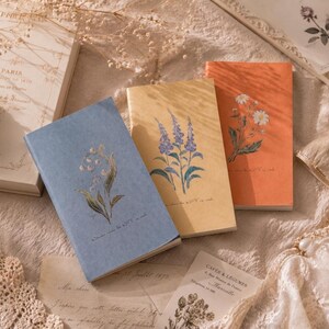 May include: Three rectangular notebooks with textured covers in blue, yellow, and orange. Each notebook features a botanical illustration: lily of the valley on the blue, lavender on the yellow, and daisies on the orange. Small cursive text is printed below each floral design.