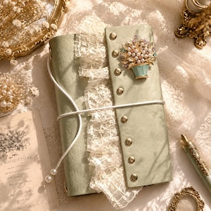 May include: A sage green journal featuring a vertical strip of white lace trim, a white cord closure with two pearl beads, and a gold-toned metal brooch shaped like a flower basket filled with pearls and crystals, secured by seven gold-toned studs.
