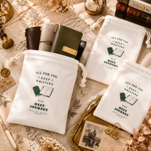 May include: Three white cotton drawstring pouches featuring the dark green printed text ALL FOR YOU KEEP WRITTING WITH BEST JOURNEY BRING YOU THE BEST FOREVER and a graphic illustration of an open notebook and a closed journal.