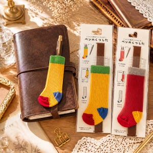 May include: Two sock-shaped pen holders on cardboard backing cards, one yellow with a green cuff and red and blue toe, the other red with a grey cuff and yellow toe. A third yellow sock pen holder is attached to a brown leather notebook.