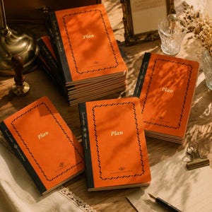 May include: A collection of rectangular notebooks with bright orange covers and black spines. Each cover features a decorative black rope-style border, the word "Plan" printed in the center, and the text "Travel notes" along the left edge.