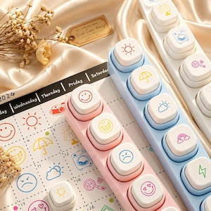 Può includere: Tre set di timbri rettangolari in plastica rosa, azzurro e bianco, ciascuno con una fila di pulsanti quadrati con icone colorate tra cui faccine sorridenti, simboli meteorologici e oggetti, appoggiati su una pagina di calendario 2024.