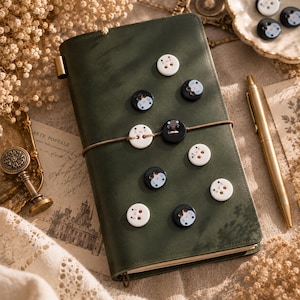 May include: A dark green leather-bound journal secured with a thin elastic band, featuring several small, round buttons decorated with cat face illustrations in black and white, arranged across the front cover surface.