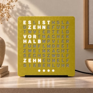 Könnte beinhalten: Eine quadratische, senfgelbe Wortuhr mit einem Buchstabenraster. Der beleuchtete weiße Text lautet ES IST ZEHN VOR HALB ZEHN. Vier kleine weiße, kreisförmige Lichter befinden sich unten in der Mitte des Zifferblatts mit dezentem geometrischem Muster.