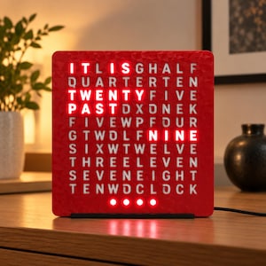 Peut inclure: Une horloge à mots carrée rouge avec une surface texturée présentant une grille de lettres blanches. Le texte rouge illuminé indique l'heure IT IS TWENTY PAST NINE. Quatre petits voyants circulaires rouges sont situés au bas du cadran.