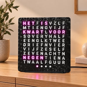 Op de afbeelding: Een vierkante woordklok met een zwart, getextureerd oppervlak en een raster van witte letters. De woorden HET, IS, KWART, VOOR en NEGEN zijn felroze verlicht en geven de tijd aan. Onderaan bevinden zich vier kleine ronde lichtpuntjes.