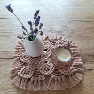 Könnte beinhalten: Ein quadratischer Makramee-Untersetzer in hellem Beige mit geknotetem Muster und Fransen an den Rändern. Eine kleine weiße Keramikvase mit violetten Lavendelzweigen und ein rundes Glas-Teelichtglas mit einer weißen Kerze stehen auf dem Untersetzer.