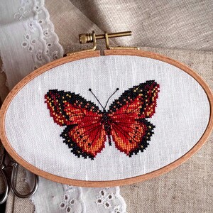 May include: An oval wooden embroidery hoop containing white fabric with a cross-stitch design of a butterfly. The butterfly features vibrant orange and red wings with detailed black outlines and patterns. A metal tension screw is at the top.
