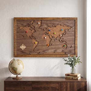 May include: A rectangular wooden world map wall art piece featuring a cork inlay with laser-cut continents and country borders. The map includes a decorative compass rose with text and several colorful push pins inserted into various locations.