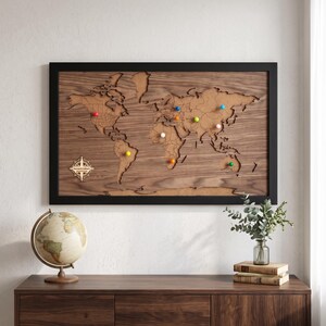 May include: A rectangular wall-mounted world map featuring a dark wood frame and a layered wooden surface with engraved country borders. The map includes a compass rose design and several colorful push pins placed across various continents.