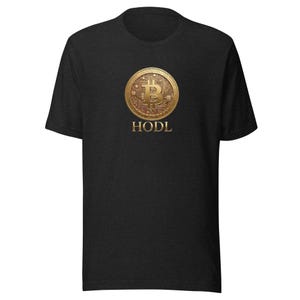 Puede incluir: Una camiseta de manga corta en color gris carbón con un gráfico central de una moneda Bitcoin de oro metálico con detalles de circuitos. Debajo de la moneda, la palabra HODL está impresa en una fuente serif dorada a juego.
