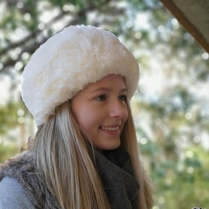 May include: A white faux fur headband with a wide band and a fluffy texture. The headband is worn by a person with long blonde hair. The background is blurred and out of focus.