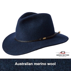 May include: A navy blue felt fedora hat with a black braided band and a gold metal accent. The hat is made of Australian merino wool. "Grizzly Hatters Australia" is printed on the hat's band.