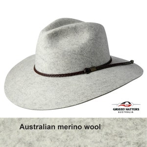 Op de afbeelding: Een grijze wollen fedora hoed met een bruin leren gevlochten band en een metalen gesp. De hoed is gemaakt van Australische merinowol. De hoed is schuin gezet en de rand is breed. De hoed is gemaakt door Grizzly Hatters Australia.