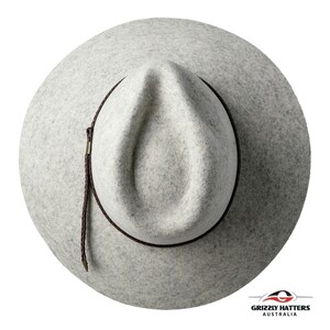 Op de afbeelding: Een lichtgrijze wollen fedora-hoed met een bruin leren band en een bruin gevlochten koord. De hoed heeft een licht ingedeukte kroon en een brede rand.