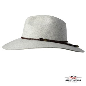 Op de afbeelding: Een lichtgrijze wollen fedora-hoed met een gevlochten band van bruin leer en een klein metalen accent. De hoed heeft een brede rand en een licht ingedeukte kroon.