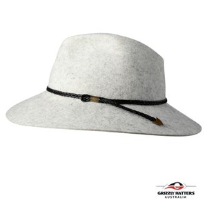 Op de afbeelding: Een lichtgrijze wollen fedora-hoed met een zwarte gevlochten band en een gouden metalen accent. De hoed heeft een brede rand en een lang zwart koord.