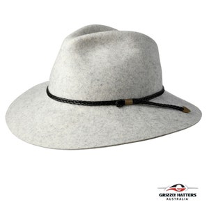 Op de afbeelding: Een lichtgrijze wollen fedora-hoed met een zwarte gevlochten leren band en een gouden metalen accent. De hoed heeft een brede rand en een licht ingedeukte kroon.
