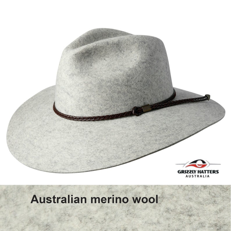 Op de afbeelding: Een grijze wollen fedora hoed met een bruin leren gevlochten band en een metalen gesp. De hoed is gemaakt van Australische merinowol. De hoed is schuin gezet en de rand is breed. De hoed is gemaakt door Grizzly Hatters Australia.