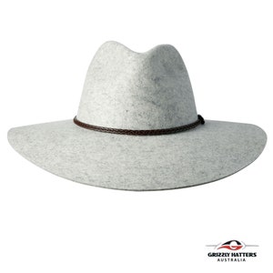 Op de afbeelding: Een lichtgrijze wollen fedora hoed met een bruin leren band. De hoed heeft een brede rand en een licht ingedeukte kroon.