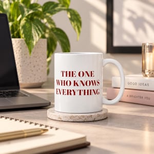 Könnte beinhalten: Ein weißer Keramikbecher mit Henkel, auf dessen Vorderseite der rote Text "THE ONE WHO KNOWS EVERYTHING" aufgedruckt ist. Der Becher steht auf einem runden, gesprenkelten, hellen Untersetzer aus Stein auf einer Schreibtischoberfläche.