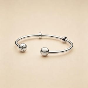 May include: A polished silver-tone open cuff bracelet featuring two large spherical end caps and two smaller sliding spherical beads along the band. The larger end caps are engraved with the text PANDORA, showcasing a sleek and reflective metallic finish.