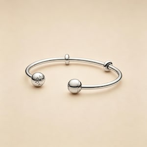 May include: A polished silver-tone open cuff bracelet featuring two large spherical end caps and two smaller cylindrical spacers along the band. The spherical end caps are engraved with the text PANDORA, featuring a small symbol above the letter O.