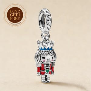 May include: A silver dangle charm shaped like a nutcracker soldier, featuring a blue crown, red jacket with black details, and a green heart emblem on the chest. The charm has a bail at the top with engraved text and a polished metallic finish.