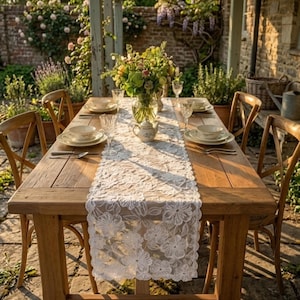 Könnte beinhalten: Ein weißer Tischläufer aus Spitze mit Blumenmuster auf einem rechteckigen Holztisch. Der Tisch ist mit weißen Keramikschalen, klaren Weingläsern und einer kleinen weißen Teekanne gedeckt, in deren Mitte ein großer Strauß grüner Blumen steht.