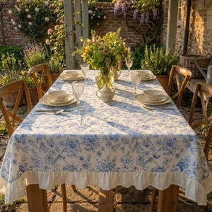 May include: A rectangular dining table covered with a white tablecloth featuring a blue floral pattern and a ruffled hem. The table is set with white ceramic plates, clear glass goblets, and a central glass vase filled with a bouquet of flowers.