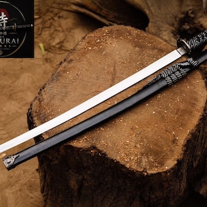 May include: A katana sword with a polished silver blade and a black handle wrapped in white cord. The black scabbard features a matching white cord wrap and silver metal accents. A logo in the corner displays the text The Samurai Works.