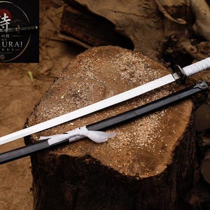 Puede incluir: Una espada katana con una hoja larga, recta y de color plateado, y un mango envuelto en blanco. La espada viene acompañada de una vaina negra con una cinta blanca. Un logotipo en la esquina muestra el texto The Samurai Works.