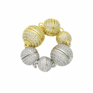Op de afbeelding: Een cirkelvormige sieraadsluiting bestaande uit vijf bolvormige onderdelen verbonden door kleine metalen ringetjes. De bollen zijn ingelegd met heldere pavé-steentjes en hebben een combinatie van goudkleurige en zilverkleurige metalen afwerkingen.