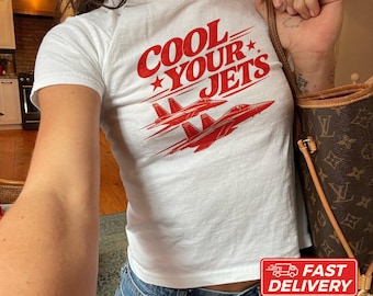 May include: A white short-sleeved t-shirt featuring red text that reads COOL YOUR JETS with a graphic of two red fighter jets flying in formation. The shirt is made of cotton and is shown in a size XS. The design includes small red stars.