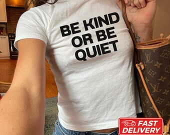 May include: A white short-sleeved t-shirt featuring the black text BE KIND OR BE QUIET printed on the front. The shirt is paired with blue denim jeans and a brown patterned shoulder bag with gold-toned hardware accents.