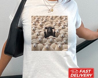 May include: A white short-sleeve t-shirt featuring a central graphic print of a large flock of white sheep surrounding a single black sheep in the center that is raising both of its middle fingers toward the viewer.