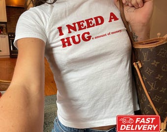 May include: A white short-sleeved t-shirt featuring red text that reads I NEED A HUG e amount of money. The shirt is paired with a brown patterned shoulder bag and blue denim jeans. The garment is made of cotton and is shown in a size XS.