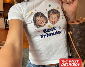 May include: A white short-sleeve t-shirt featuring a graphic print of two children's faces, colorful stars, and the text "Best Friends" in dark blue lettering. The shirt is made of 100% cotton and is shown in size XS.