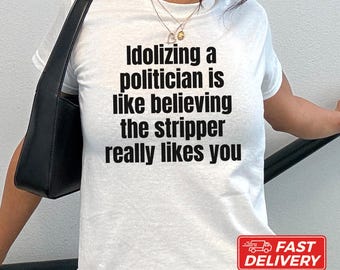 May include: A white short-sleeved t-shirt featuring black text that reads: Idolizing a politician is like believing the stripper really likes you. The shirt is made of 100% US cotton and the model is wearing a size XS for a standard fit.