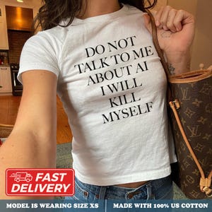 May include: A white short-sleeved t-shirt featuring black text that reads DO NOT TALK TO ME ABOUT AI I WILL KILL MYSELF. The shirt is made of 100% US cotton and is shown in size XS. A brown patterned handbag is visible on the right side of the image.
