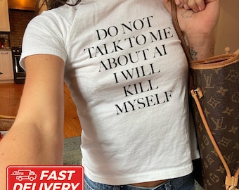 May include: A white short-sleeved t-shirt featuring black text that reads DO NOT TALK TO ME ABOUT AI I WILL KILL MYSELF. The shirt is made of 100% US cotton and is shown in size XS. A brown patterned handbag is visible on the right side of the image.