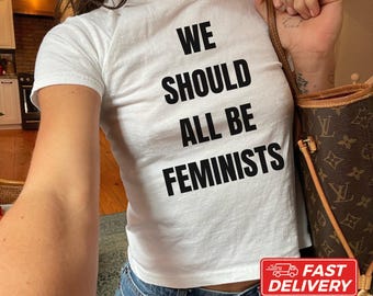 May include: A white short-sleeved t-shirt featuring the black text WE SHOULD ALL BE FEMINISTS printed on the front. The shirt is paired with blue denim jeans and a brown patterned shoulder bag with gold-toned hardware accents.